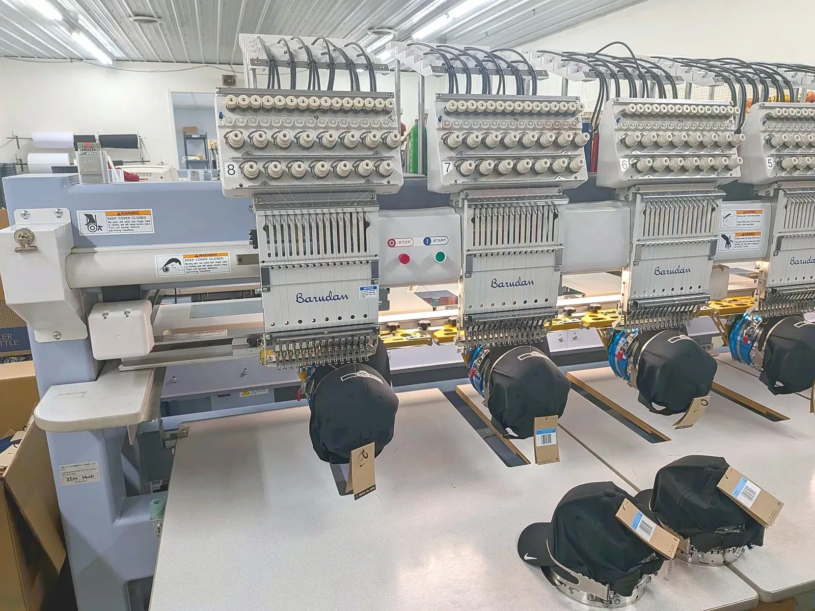 A row of multi-head embroidery machines stitching black caps in Del Print's embroidery department
