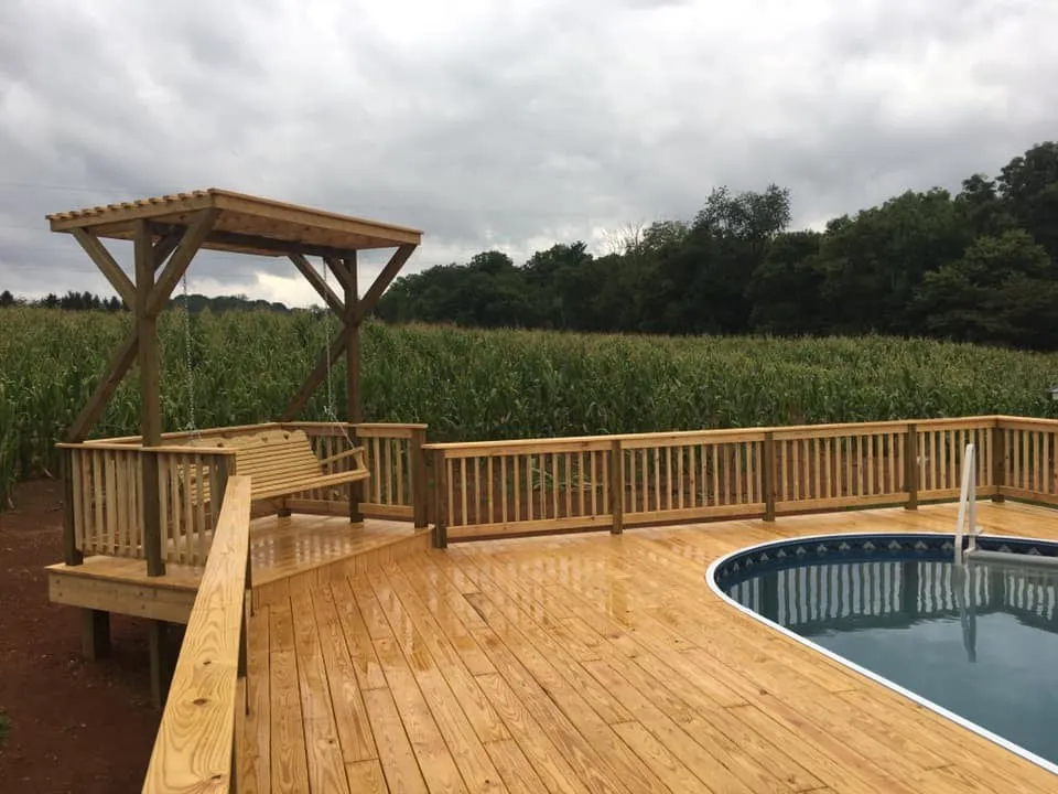 A wood pool deck with built-in bench seating, railings and a pergola, beside a cornfield