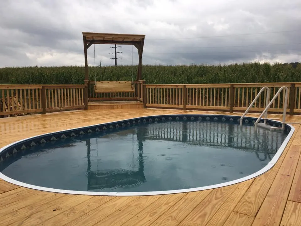 A wide pressure-treated wood deck built around an above-ground pool, with a pergola swing at the far railing and a cornfield beyond